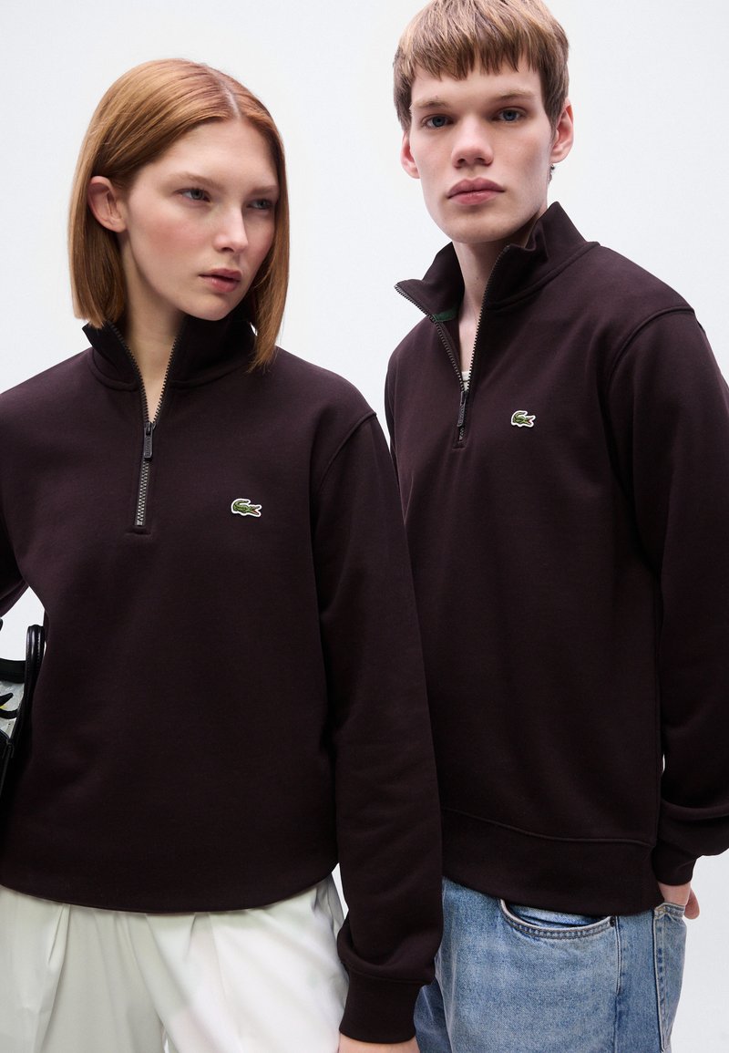 Pullovers marron foncé avec col à demi-fermeture éclair, arborant un logo vert subtilement brodé sur la poitrine et des poignets côtelés.