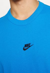 Ljusblå bomullströja med rund hals och svart broderad Nike-logga på vänster bröst. Slät textur, avslappnad design.