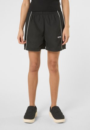 Osoba ubrana w czarne szorty z elastycznym pasem, białym paskiem po bokach i logo FILA, w połączeniu z czarnymi wsuwanymi butami na białej podeszwie.