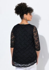 Top in pizzo nero con maniche a tre quarti, caratterizzato da un intricato motivo floreale, un colletto tondo e un orlo posteriore più lungo.