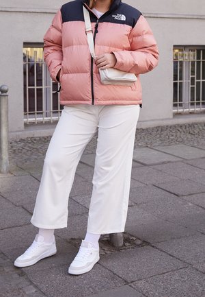 Person iført en pink og sort North Face-jakke, hvide vidde bukser, hvide sneakers og bærende en lille beige crossbody taske står på en fortov.