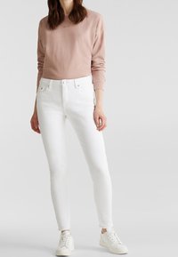 Vaaleanpunainen pitkähihaisen huppari, jossa on brodeerattu logo, yhdistettynä valkoisiin slim fit -farkkuihin ja valkoisiin lenkkareihin. Pehmeä tekstuuri, vartalonmyötäinen muotoilu.