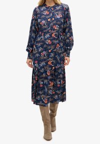 Abito a maniche lunghe con stampa floreale in blu navy con accenti arancioni e rosa, design a strati e orlo fino al ginocchio. Indossato con stivali taupe alti fin sopra il ginocchio.
