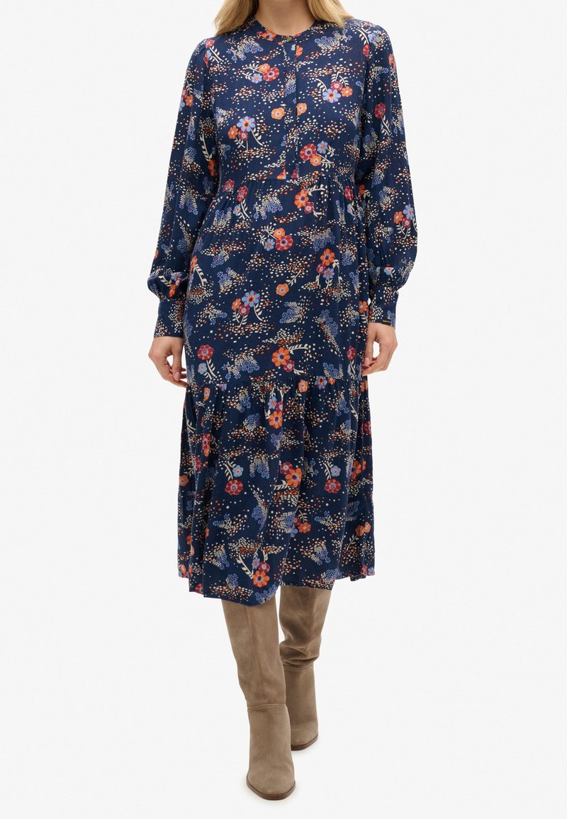 Abito a maniche lunghe con stampa floreale in blu navy con accenti arancioni e rosa, design a strati e orlo fino al ginocchio. Indossato con stivali taupe alti fin sopra il ginocchio.