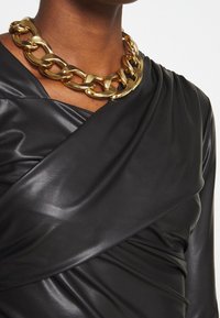 Schwarzes Kunstleder-Wickeltop mit drapiertem Design; kombiniert mit einer massiven Goldkette mit großen Gliedern und glänzender Oberfläche.