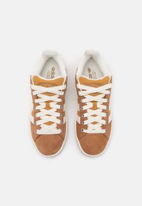 Scarpe da ginnastica marroni e gialle in materiali scamosciati e in tessuto, con lacci bianchi e tre strisce bianche sui lati.