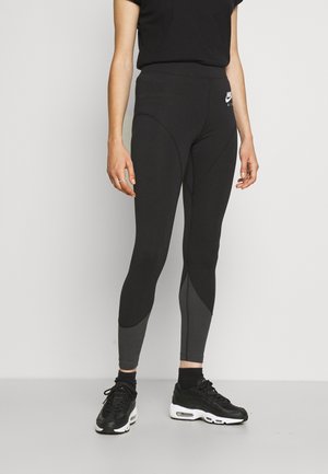 Persona vistiendo leggings Nike Air en negro y gris oscuro con zapatillas negras, de pie contra un fondo liso gris claro.