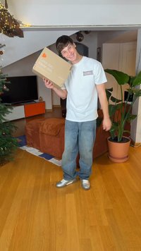 Boîte en carton avec le texte « Aime-moi, Porte-moi » tenue par une personne portant un t-shirt blanc et un jean large bleu, à l'intérieur avec des plantes et du mobilier.