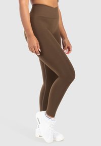 Leggings marroni a vita alta realizzati in tessuto elastico, con una texture liscia e un dettaglio di cucitura laterale, abbinati a scarpe da ginnastica bianche.