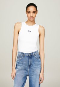Top sin mangas blanco, ajustado, con cuello redondo, que presenta "Tommy" bordado en azul marino. Combinado con vaqueros de denim azul de cintura alta.
