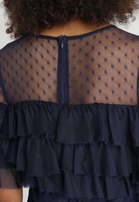 Robe bleu marine avec un corsage en mesh transparent à texture à pois, détails à volants superposés et fermeture éclair au dos.
