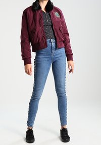 Person som bär en vinröd bomberjacka med svart pälskrage, mörkt mönstrad skjorta, höga skinny jeans med snörning på sidan, och svarta sneakers.