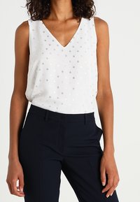 Haut sans manches blanc avec un col en V, agrémenté de bosses argentées. Associé à un pantalon ajusté bleu marine. Texture de tissu lisse.