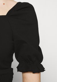 Black top with puffed short sleeves, featuring a gathered ruffle detail at the cuffs and a fitted waistline. Smooth cotton fabric.