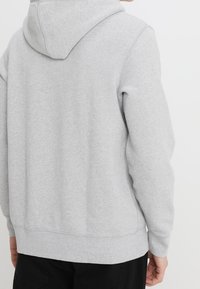 Persona vistiendo una sudadera gris claro vista desde la espalda, combinada con pantalones negros frente a un fondo blanco.