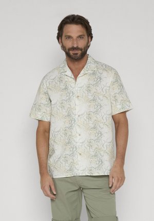 Hombre con barba vistiendo una camisa de manga corta de color claro con patrones abstractos y pantalones verde oliva sobre un fondo liso.