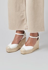 Sandalias de cuña espadrille blancas con punta cerrada, que presentan una tapa beige, una correa para el tobillo y una suela de cuerda de yute natural.