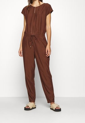 Kobieta ubrana w brązowy kombinezon z krótkim rękawem z regulowanym paskiem w talii i beżowe sandały na platformie bez pasków, stojąca na tle jednolitej białej ściany.