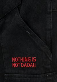 Schwarzer Stoff mit strukturiertem Finish, mit rotem, gesticktem Text auf der vorderen Tasche, der "NICHTS IST NICHT DADA!!" liest.