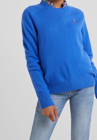 Blå stickad tröja med räfflad halsringning och muddar, tillverkad av ett mjukt material, med en liten broderad logotyp på bröstet.