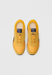 Dzelteni sporta apavi ar gludu sintētisku virsu, mīkstinātu apkakli, teksturētām auklām un gumijas zoli. Uz mēles ir zils Reebok logo.