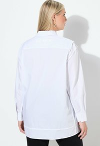 Witte button-up shirt met een ontspannen pasvorm, voorzien van een kraag en zwarte tekst "BLACK" op de achterkant, benadrukt door lange mouwen en een afgeronde zoom.