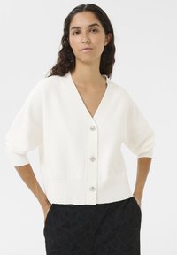 White knitted cardigan with a deep V-neck, featuring four decorative button closures and three-quarter sleeves. Worn with textured black pants.
