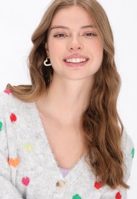 Cárdigan gris de punto con patrones de corazones coloridos, cuello redondo y cierre con un solo botón. El accesorio destacado es un pendiente en forma de corazón.