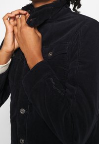 Marinblå kordjacka med ställningkrage, tryckknappar, två bröstfickor och elastiska ärmslut. Tygets textur är tydligt ribbad.