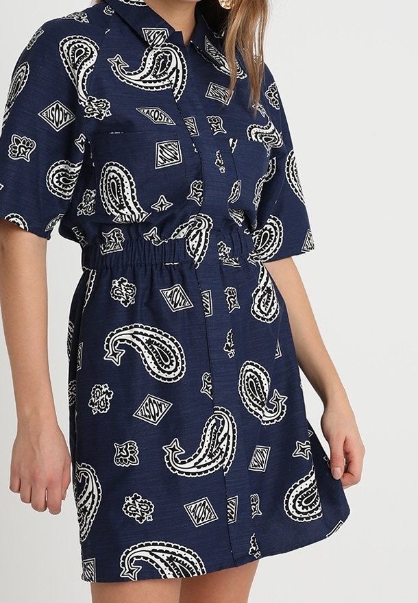 Femme portant une robe bleu marine à manches courtes avec des motifs paisley blancs et géométriques et une taille élastique.