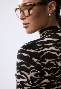 Haut à manches longues rayé noir et beige avec un col montant, une texture côtelée et une coupe ajustée. Grandes lunettes écaille de tortue et boucles d'oreilles goutte en or.