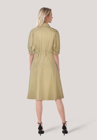 Knie kurzer, olivgrüner Kleid mit Taillengurt, Puffärmeln und glatter Textur. Getragen mit schwarzen High-Heels.