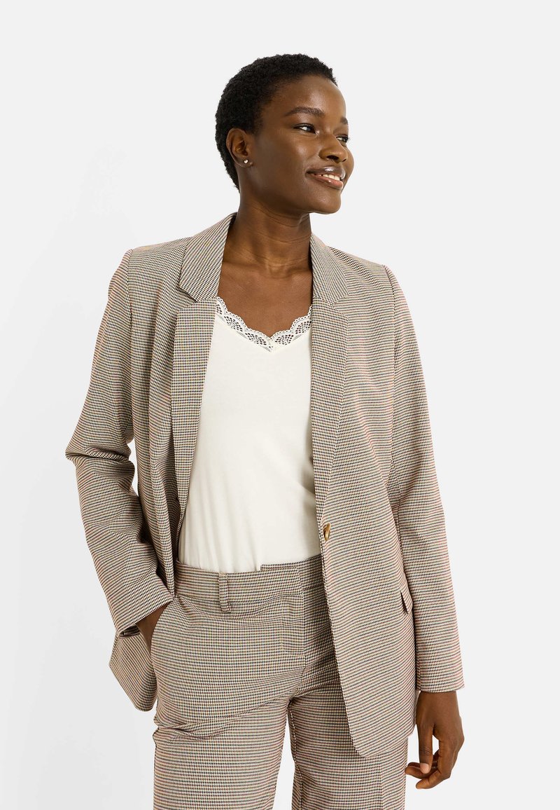 Blazer à carreaux marron et crème avec un seul bouton, porté sur un haut à dentelle blanche, assorti à un pantalon à motif similaire.