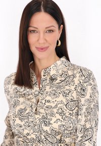 Bluzka z kołnierzykiem w odcieniu kremowym z czarnym kwiatowym wzorem paisley; detale z guzikami i miękka tkanina. Modelka nosi złote kolczyki w kształcie kół.