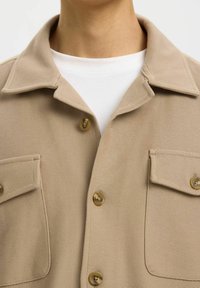 Beige textuur jack met knoopzakken op de borst gedragen over een wit T-shirt met ronde hals, waarbij de kraag en het bovenste deel van de borst zichtbaar zijn.