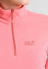 Różowy polarowy rozpinany golf z wysokim kołnierzem i półzamkiem. Zawiera haftowane logo w dopasowanym odcieniu różu oraz gładką fakturę.