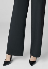 Kobieta w czarnych szerokich spodniach i czarnych szpilkach na lekkoszarym tle.