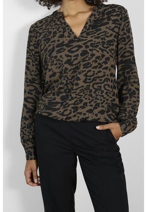 Vrouw die een lange mouws blouse draagt met een bruin-zwarte luipaardprint en een V-hals, gecombineerd met een zwarte broek, één hand in de zak, beeld is bij de schouders afgeknipt.