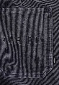Tissu de velours côtelé gris foncé présentant des côtes verticales, avec une poche en relief et une étiquette logo discrète sur le côté. Apparence texturée et douce.