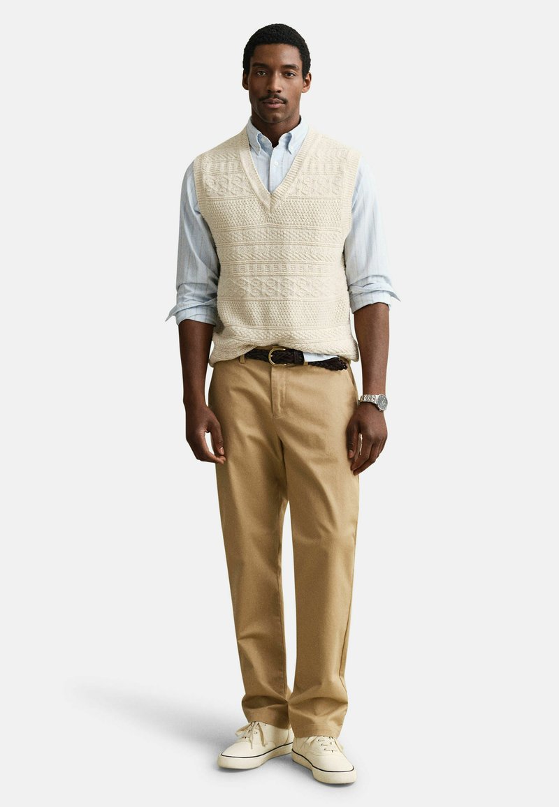 Hombre con camisa de botones azul claro, chaleco de punto trenzado crema, pantalones color beige, zapatillas blancas y reloj de pulsera, de pie mirando hacia adelante.