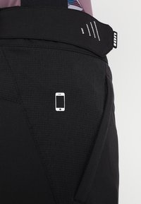 Musta ulkoiluhousut, joissa on teksturoitu kangas ja pieni valkoinen älypuhelinikoni yläreidessä vyölenkin läheisyydessä, jossa on logotaulukko.