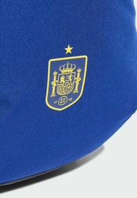 Bolsa de tela azul con un escudo amarillo con una corona, detalles intrincados en el emblema y una estrella arriba, todo sobre un fondo de textura suave.