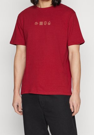 Osoba v červeném tričku se čtyřmi malými béžovými ikonami — šipka, vlny, zeměkoule, plamen — umístěnými uprostřed na hrudi, v kombinaci s černými kalhotami.