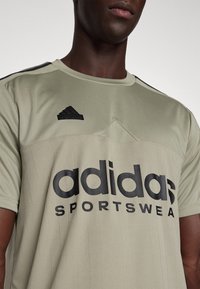 Khakifärgad sport-t-shirt med texturerat tyg, svarta vertikala ränder och stor svart "adidas"-logotyp. Har rund halsringning och korta ärmar.