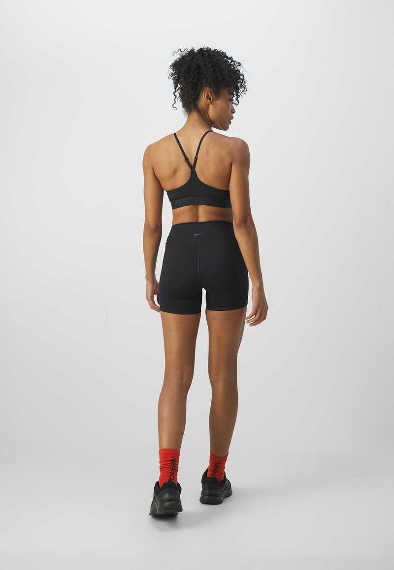 Zwarte sportbeha met dunne bandjes, gecombineerd met hoge zwarte shorts. Het model draagt zwarte schoenen en felrode sokken.