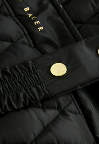 Tissu matelassé noir avec une finition brillante, comportant un bouton doré, un élastique froncé et un logo discret en lettres dorées.