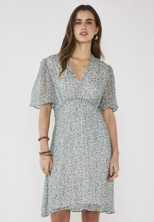 Vrouw die een lichtblauwe bloemenjurk met V-hals en korte pofmouwen draagt, gecombineerd met oorringen en meerdere armbanden.