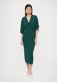 Πράσινο φόρεμα μήκους έως το γόνατο με βαθύ V λαιμόκοψη, λεπτομέρεια κόμπου στη μέση και αέρινες μανίκια. Λεία υφή, εφαρμοστός σιλουέτα.