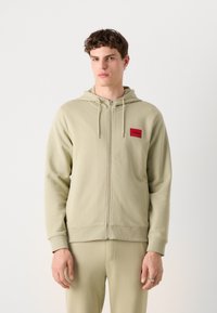 Felpa verde chiaro con zip realizzata in un tessuto morbido, caratterizzata da una toppa rettangolare rossa con la scritta "HUGO" sulla parte sinistra del petto.
