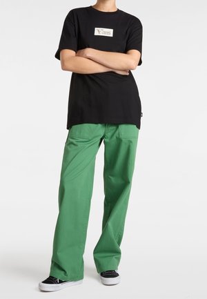 Sort bomulds t-shirt med et hvidt Vans-logo, parret med grønne vide bukser. Outfittet afsluttes med sort-hvide sneakers.